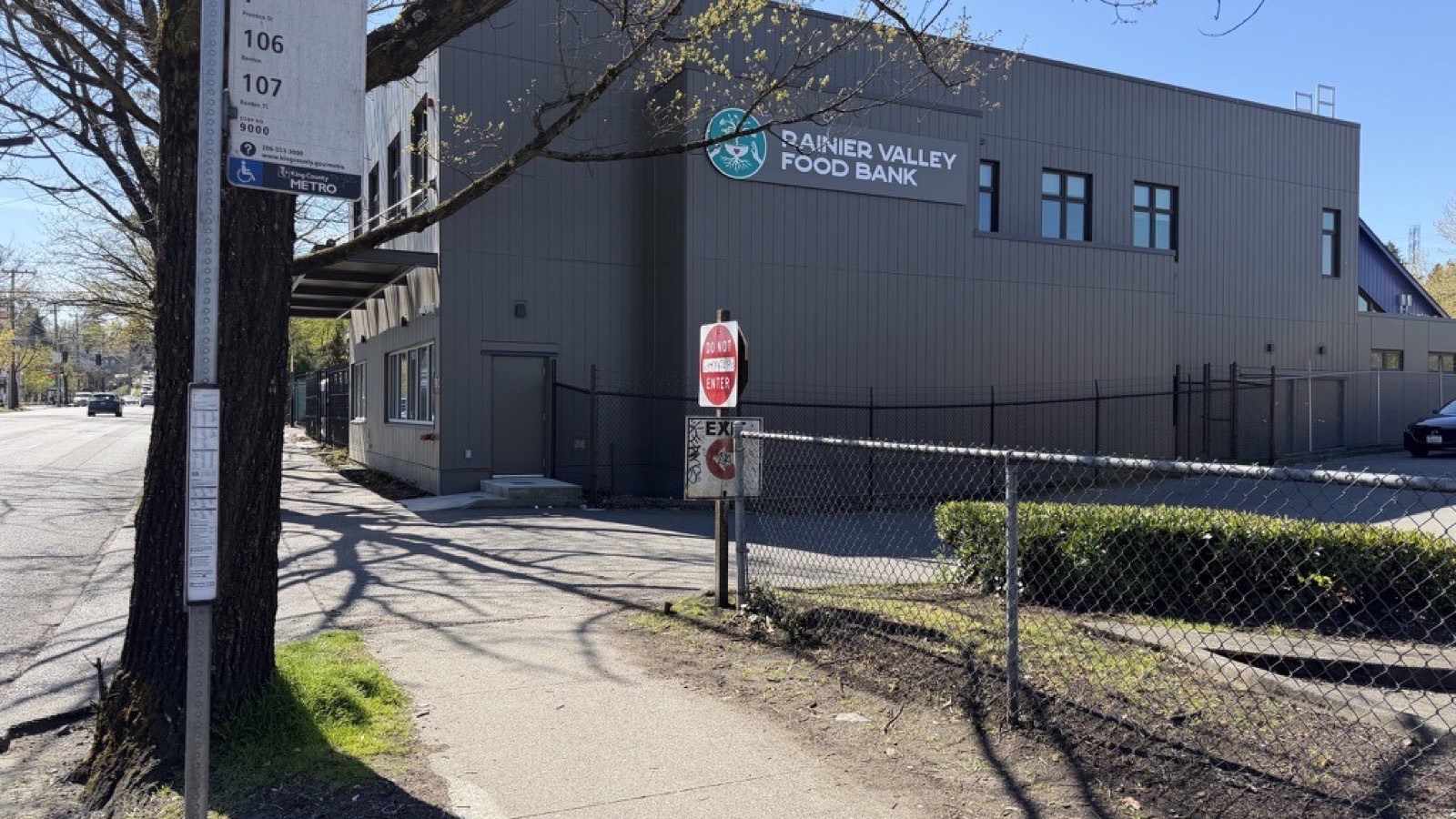 The Rainier Valley Food Bank building photographed from the sidewalk, with a King County Metro bus stop sign for routes 7, 106 and 107 in the foreground.
