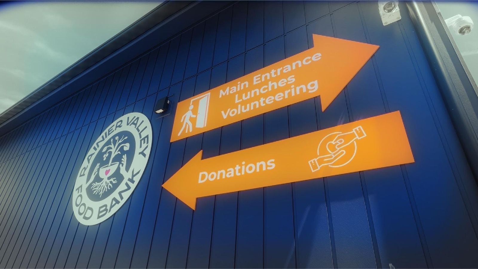 A frame from the outreach video showing the Rainier Valley Food Bank exterior with large orange arrows pointing to the main entrance, lunches, volunteering, and donations.