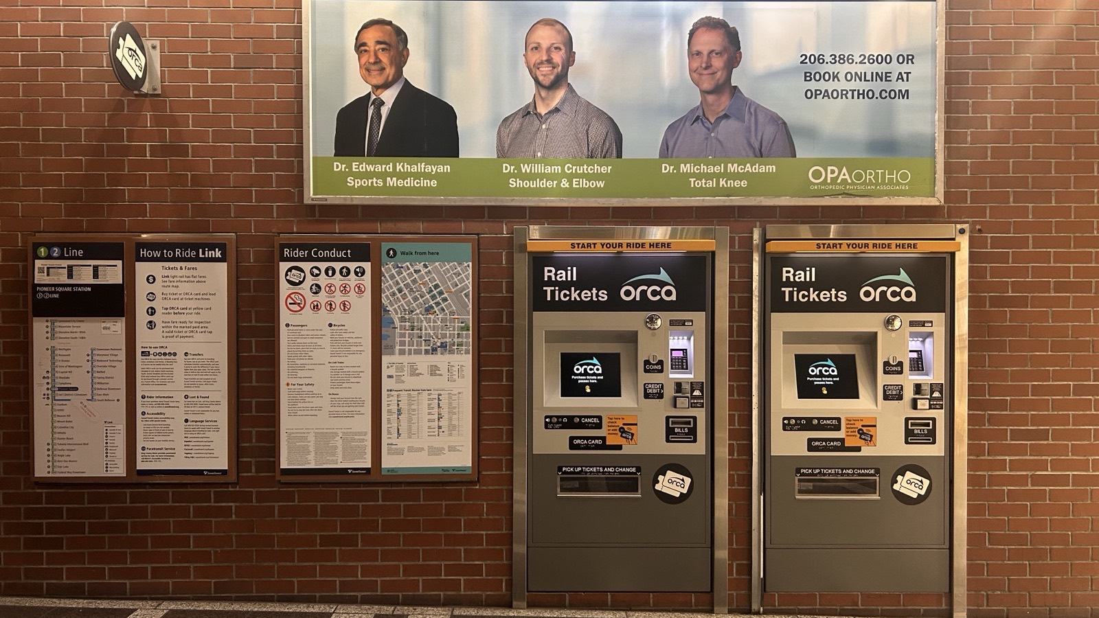 A brick wall inside Pioneer Square Station holding a Link line map, a How to Ride Link panel, a Rider Conduct panel, a local area map, and two ORCA rail ticket vending machines.